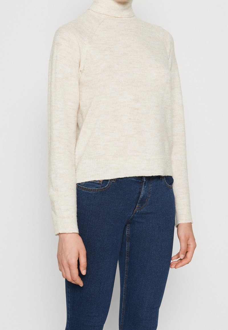 Eine Person trägt einen cremefarbenen Rollkragenpullover und dunkle blaue Jeans und steht vor einem schlichten hellen Hintergrund.