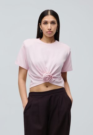 Ung kvinde med langt mørkt hår iført en lyserød cropped T-shirt med en central stofblomsterdetalje og sorte bukser med høj talje.