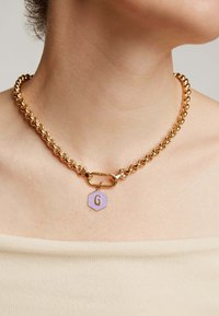 Collar de cadena de oro con grandes eslabones entrelazados, con una etiqueta hexagonal púrpura que muestra la letra "G". Textura suave y acabado brillante.