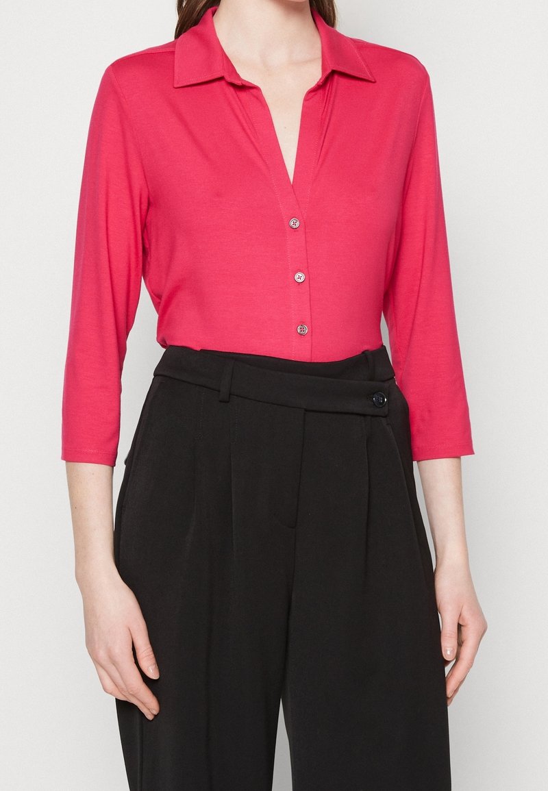 Femme portant une chemise rose vif à col avec trois boutons et un pantalon noir taille haute avec une ceinture boutonnée.