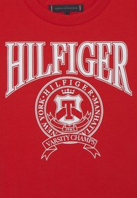 Czerwona koszula z dużym białym napisem "HILFIGER", okrągłym emblematem z literą "T", koroną, rokiem "1985" oraz wstążką "Varsity Champs" poniżej.