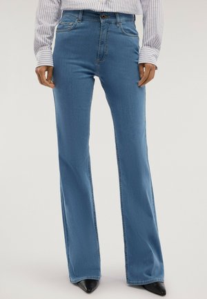 Person trägt hellblaue Bootcut-Jeans, schwarze spitze Schuhe und ein weißes Langarmhemd mit dünnen vertikalen Streifen.