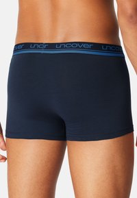 Onderrug en bovenbenen van een man, gekleed in marineblauwe boxershorts met een donkerkleurige tailleband waarop het woord "uncover" in blauw staat.