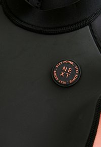 Matériau de combinaison de plongée noir avec une texture lisse, présentant un logo circulaire orange et noir portant l'inscription "RESTEZ ACTIF SUIVANT." Couture détaillée visible.