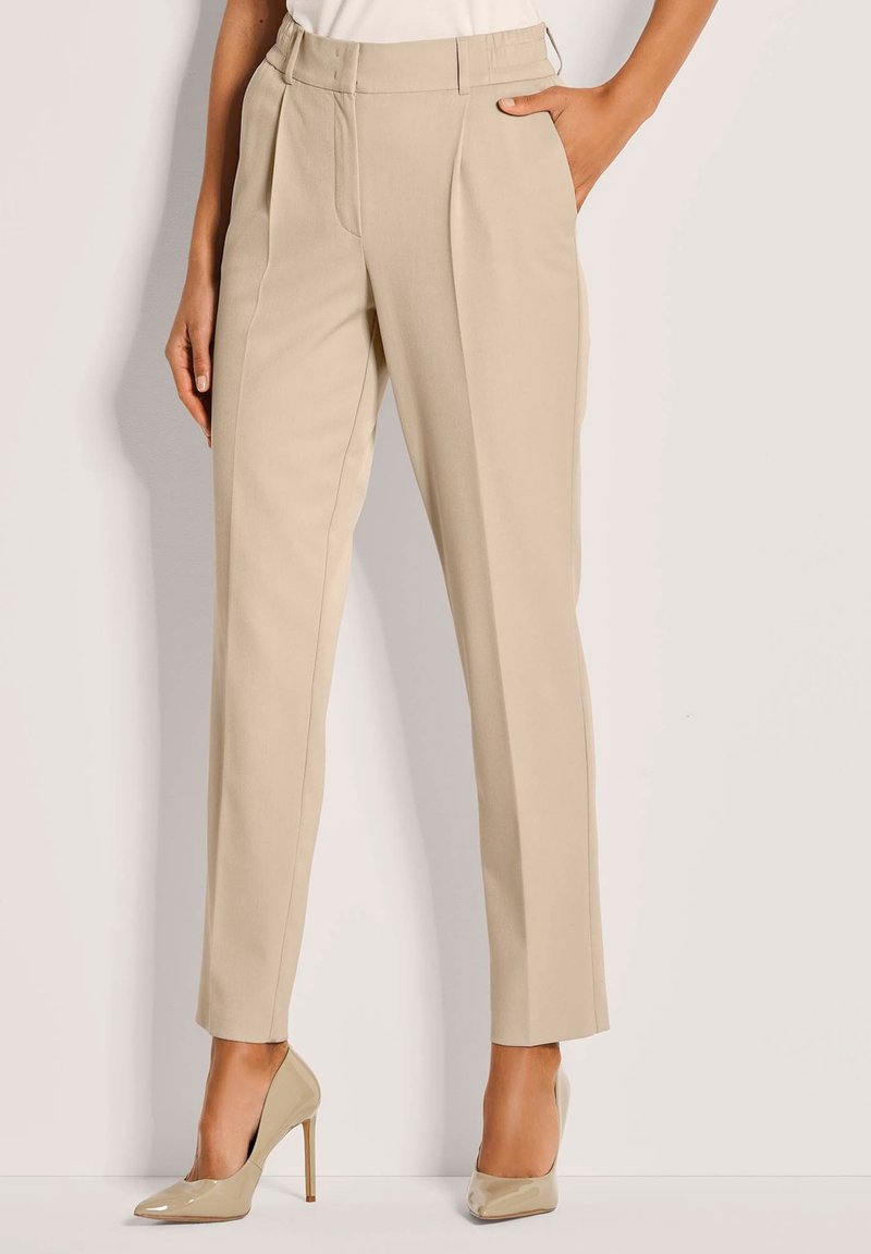 Vrouw in beige, op maat gemaakte broek en beige patenten hoge hakken, staand met één hand in de zak tegen een simpele muur.