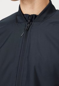 Giacca nera leggera con colletto alto, chiusura frontale con zip e texture liscia; presenta un design minimale e senza motivi visibili.
