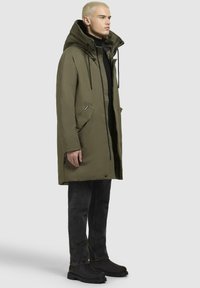 Parka verde oliva con cappuccio, zip frontale, grandi tasche laterali e tessuto testurizzato. Indossata sopra un dolcevita nero e jeans scuri.