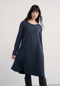 Nainen, jolla on päällä pitkähihaista tummanharmaata neulemekkoa, mustat legginsit ja mustat saappaat, seisoo vaaleaa taustaa vasten.