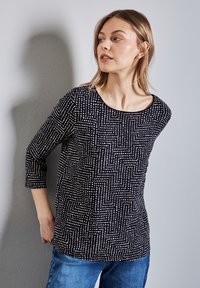 Svart topp med vitt geometriskt mönster, rundad halsringning, trekvartslånga ärmar, i kombination med ljusblå jeans, vilket visar en avslappnad design.