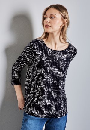 Haut noir avec un motif géométrique blanc, décolleté en scoop, manches trois-quarts, associé à un jean bleu clair, présentant un design décontracté.