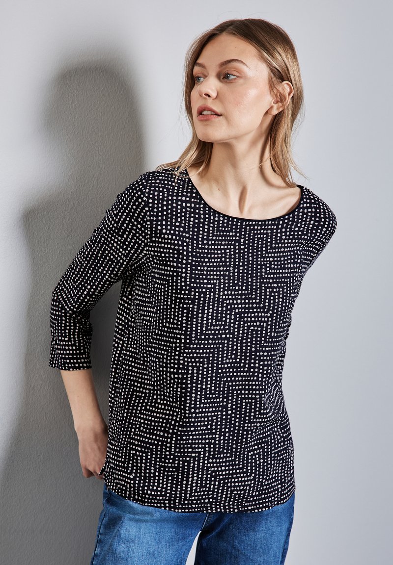 Svart topp med vitt geometriskt mönster, rundad halsringning, trekvartslånga ärmar, i kombination med ljusblå jeans, vilket visar en avslappnad design.