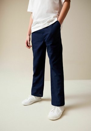 Copil purtând pantaloni bleumarin, adidași albi și un tricou alb, cu o mână în buzunar, pe un fundal simplu bej.