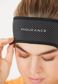 Svart atletisk pannband med vita prickar, med "ENDURANCE"-logotyp i vit text. Slät tyg och elastisk kant för passform.