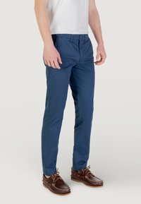 Chinos in cotone blu da uomo con vestibilità slim, dotati di chiusura con bottone, tasche laterali e una texture morbida, abbinati a scarpe da barca marroni.