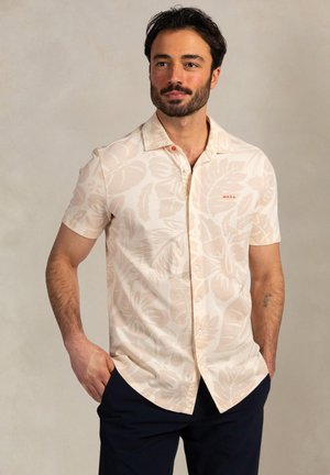 Man draagt een beige, kort mouwen, bloemmotief overhemd met knopen en donkerblauwe broek, staand met handen in de zakken tegen een effen achtergrond.