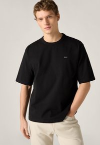 Μαύρο βαμβακερό T-shirt με στρογγυλή λαιμόκοψη και κοντά μανίκια. Φέρει ένα μικρό, τονικό λογότυπο στο στήθος. Φορεμένο με ανοιχτόχρωμο παντελόνι.