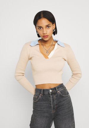 Sweater de malha canelada bege com decote em V profundo e detalhe de colarinho azul claro. Combinado com jeans escuros de cintura alta.