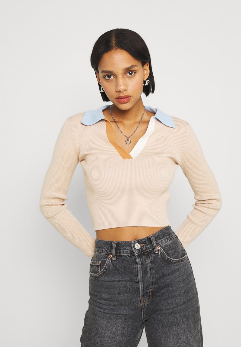 Sweater de malha canelada bege com decote em V profundo e detalhe de colarinho azul claro. Combinado com jeans escuros de cintura alta.