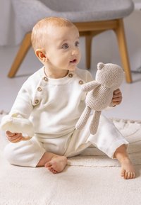Outfit bianco lavorato a maglia con bottoni, tiene in braccio un orsacchiotto di cotone beige e un piccolo giocattolo color crema, seduta su un tappeto chiaro e texturizzato.