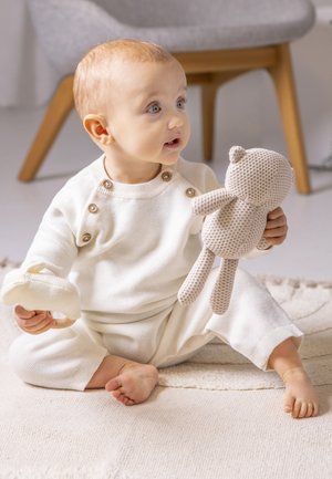 Weißes gestricktes Outfit mit Knöpfen, hält einen beigefarbenen gehäkelten Teddybären und ein kleines cremefarbenes Spielzeug, sitzt auf einem strukturierten hellen Teppich.