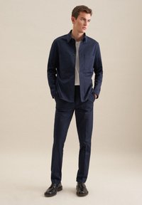 Camicia blu navy con bottoni, dotata di due tasche sul petto, abbinata a pantaloni blu navy. L'outfit presenta un design minimalista e una trama del tessuto liscia.