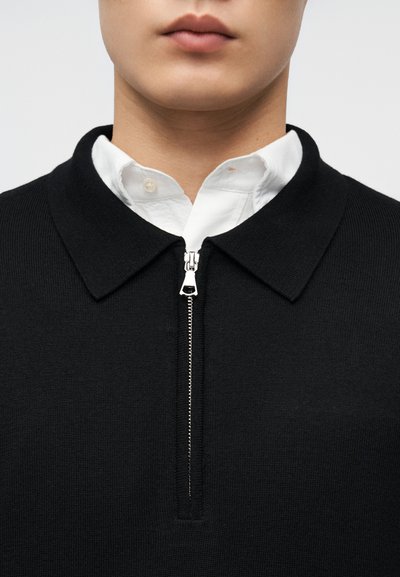 Κοντινό πλάνο ενός ατόμου που φοράει λευκό πουκάμισο με γιακά κάτω από μαύρο πουλόβερ με φερμουάρ, με το φερμουάρ να είναι εν μέρει κλειστό.