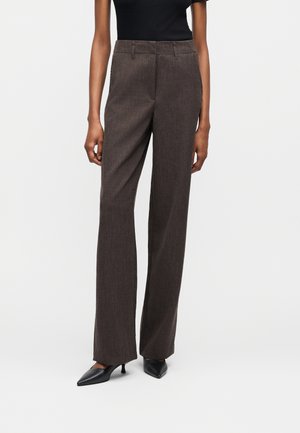 Žena nosiaca tmavohnedé nohavice s vysokým pásom a rovnými nohavicami a čierne lodičky s úzkym a špicatým päty, stojaca pred jednoliatym pozadím.