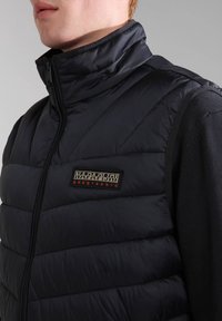 Marine blauwe gewatteerde gilet met hoge kraag, doorgestikt ontwerp en ritssluiting. Voorzien van een zwart en rood geborduurd logo op de borst. Zachte textuur.