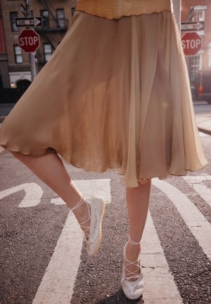 Persoon draagt een vloeiende beige rok en witte sneakers in ballerina-stijl, staand op een straat met stopborden en zebrapadmarkeringen.