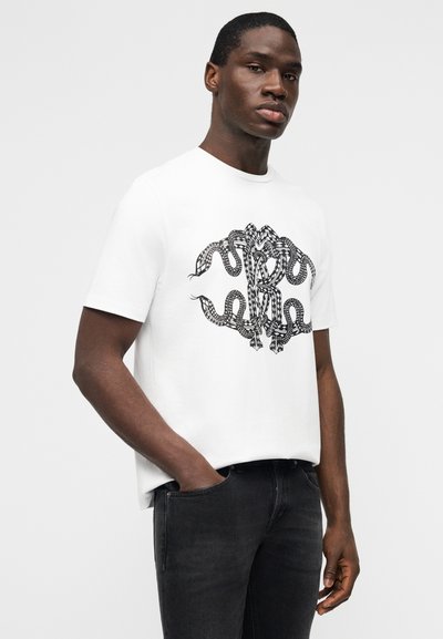 Άνδρας που φοράει άσπρο μπλουζάκι με μαύρο σχέδιο φιδιού και μαύρο τζιν, στέκεται με το ένα χέρι στην τσέπη μπροστά σε λευκό απλό φόντο.