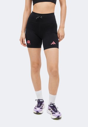 Kvinde iført sorte atletiske shorts med høj talje med lyserøde logoer, hvide ankelsokker og lilla og hvide løbesko, stående mod en ensfarvet baggrund.