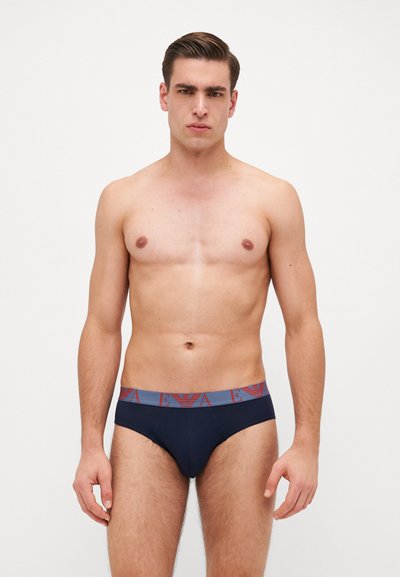 Caleçon marine avec une ceinture texturée grise ornée d'un motif de logo rouge. Le tissu semble lisse ; le design est ajusté et minimaliste.