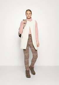 Mulher em pé, vestindo botas de caminhada bege, leggings com padrão, roupa polar rosa, colete branco sem mangas e carregando uma mochila bege num dos ombros.