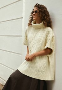 Cremfarbenes, übergroßes Strickpullover mit hohem Kragen, Zopfmuster, weiten Ärmeln, kombiniert mit einem dunkelbraunen Rock.