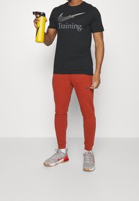 Camiseta negra de Nike con el texto "Training." pantalones jogger rojos y zapatillas grises con acentos rojos. Sostenido una botella de agua amarilla.