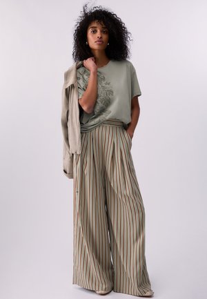 Femeie cu păr creț poartă un tricou verde cu imprimeu grafic, pantaloni largi cu dungi și ține o jachetă bej peste umăr.