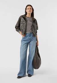 Brązowa skórzana kurtka bomberka, paskowana koszula, szerokie niebieskie dżinsy, błyszczące czarne buty oraz teksturowana brązowa torba. Neutralne tło.