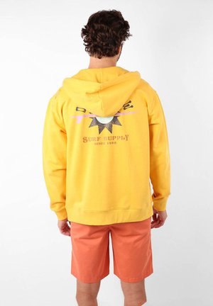 Sweatshirt à capuche jaune avec un imprimé graphique dans le dos, présentant un design de soleil et le texte "SURF SUPPLY SINCE 1985." Porté avec un short orange.