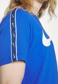 Sininen lyhythihainen t-paita, joka on valmistettu puuvillasta. Paitassa on suuri valkoinen Nike-logo ja sivusuunnassa kapea raita, jossa on toistuvia pieniä valkoisia logoja mustalla taustalla.