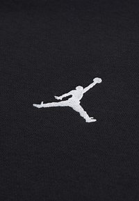 Tessuto nero con una superficie testurizzata che presenta un logo Jumpman ricamato in bianco in posizione di salto, mentre tiene in mano un pallone da basket.