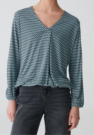 Vrouw draagt een lang mouw blauw gebloemde V-hals blouse, ingestopt in een zwarte jeans met hoge taille, staand tegen een effen lichte achtergrond.