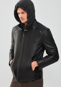 Veste en cuir noire avec une finition texturée, dotée d'une capuche grise, d'une fermeture éclair à l'avant et de poches latérales. Détails de couture sur les épaules.
