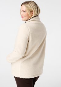 Vrouw met blond haar in een crèmekleurige sherpa fleece jas en donkere broek, iets naar links gekeerd met handen in de zakken.