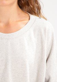 Světle šedá mikina s žebrovaným kulatým výstřihem, s měkkou texturou a knoflíkovými detaily na rameni. Tkanina vypadá ležérně a pohodlně.
