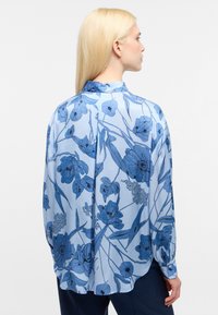 Blusa azzurro chiaro con motivo floreale in diverse tonalità di blu, maniche lunghe e chiusura a bottone al polsino. Tessuto morbido.