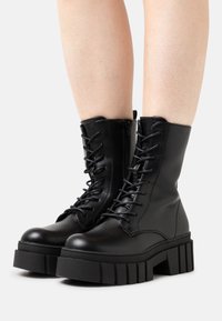 Bottes de combat en cuir noir avec semelle épaisse et striée, devant à lacets et détail de zip sur le côté, dotées d'un bout rond et d'un design robuste.