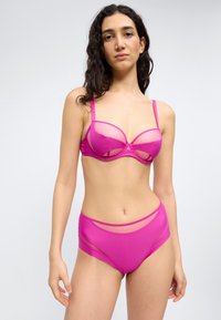 Γυναίκα με μακριά σκουρόχρωμα μαλλιά, που φορά διαφανές, φωτεινό ροζ σετ εσωρούχων, μπροστά σε απλό ανοιχτό γκρι φόντο, κοιτάζοντας προς τα εμπρός.