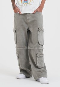 Cargo pants in olive green cotton, featuring multiple large side pockets, a relaxed fit, and straight legs. Paired with white sneakers.