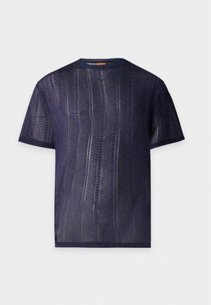 Grand homme debout, portant une chemise sombre à manches courtes avec un motif, un short noir jusqu'aux genoux et des chaussures noires à lacets sur un fond uni.