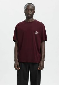 Burgundska majica s kratkimi rokavi in okrolim ovratnikom, z belim napisom "LA FLAMME ROUGE" na prsih. Tkanina je videti gladka in lahka.